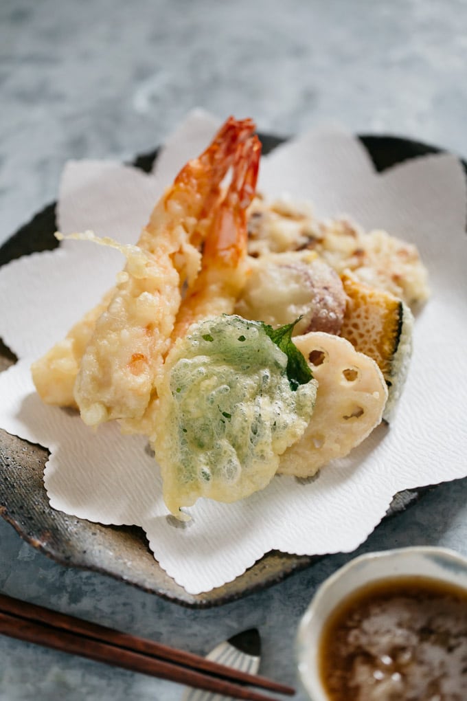 Two prawn tempura, shiso leaf, renkon, kabocha and chikuwa fish cake tempura on a plate with a bowl of tentsuyu with gated daikon in it