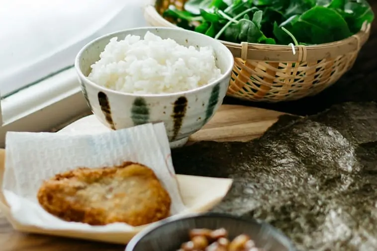 a cup of rice, spinach in a bamboo bowl, a piece of tonkatsu and nori sheet on a work surface 