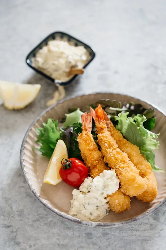 Three fried shrimp with tartar sauce on a round plate with green salad leaves, tomato and a wedge of lemon