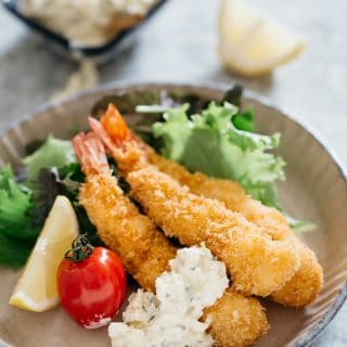 Three panko fried shrimps on a round plate with green salad, a tomato and a wedge of lemon and tartat sauce
