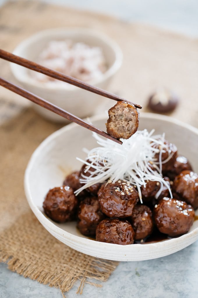 a pair of chopstick picked up a teriyaki meatball, a bowl of meatballs in background. 