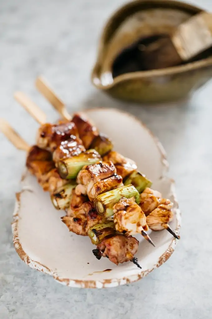 Three Yakitori skewers on Japanese pottery plate with a bowl of yakitori sauce in background