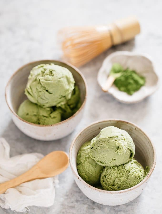 two bowls of green tea ice cream with a wooden spoon and green tea powder in a small bowl with matcha bamboo whisk in background