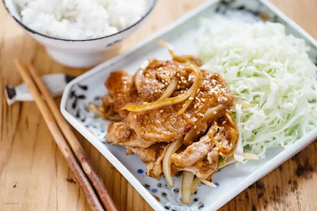 shogayaki served on a plate with shredded cabbage and a pair of chopsticks and a bowl of rice 