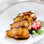 4 slices of Buri Teriyaki are served on a oval plate with a flower shaped pickled radish