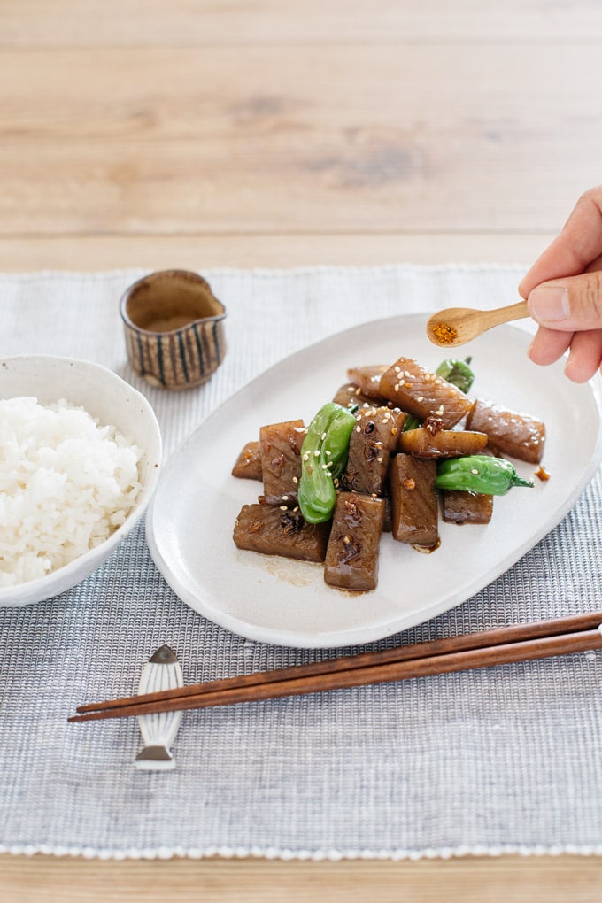 konnyaku steak with shishito peppers served on a oval shaped plate with a bowl of rice, and a pair of chopsticks and Shichimi togarashi being sprincled