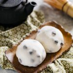 Two daifuku served on a rectangle shaped plate with a black steel teapot and a bamboo whisk.