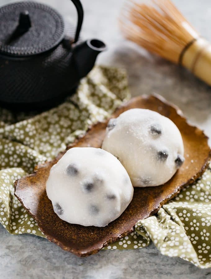 Two daifuku served on a rectangle shaped plate with a black steel teapot and a bamboo whisk.