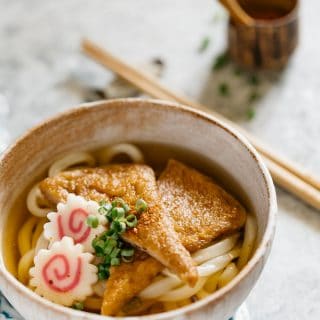 Kitsune udon served in a noodle bowl with a pair of chopstick is in background