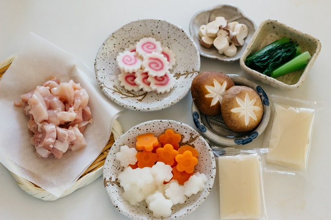 chicken chopped up on the bamboo tray, naruto slices, carrots, daikon, shiitake mushrooms, two mochi rice cakes, and Komatsuna Japanese mustard in small bowls.