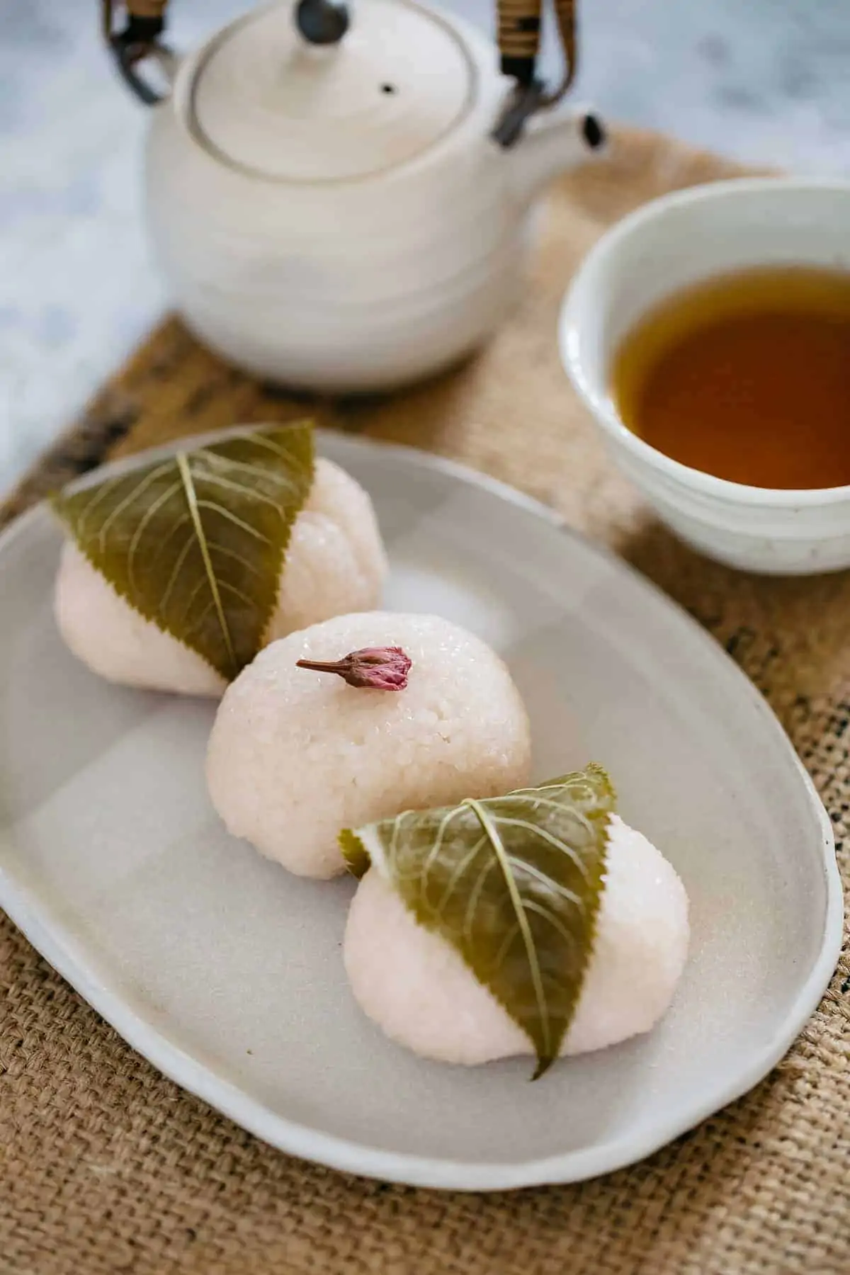 Two sakura mochi with leaves wrapped around, one sakura mochi decorated with one flower on top of it.