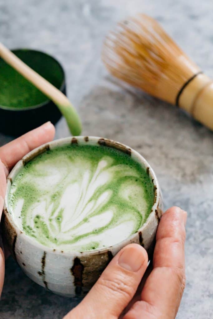 Two hands holding matcha latte bowl with a bamboo whisk in background