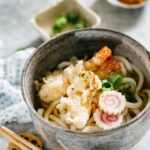 Tempura Udon served in a Udon noodle soup bowl with two shrimp tempura, two slices of Naruto and scallions.