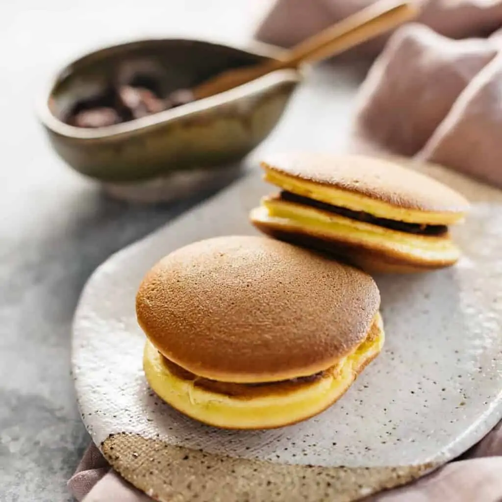  two dorayaki on a plate with sweet azuki bean paste in a bowl in background