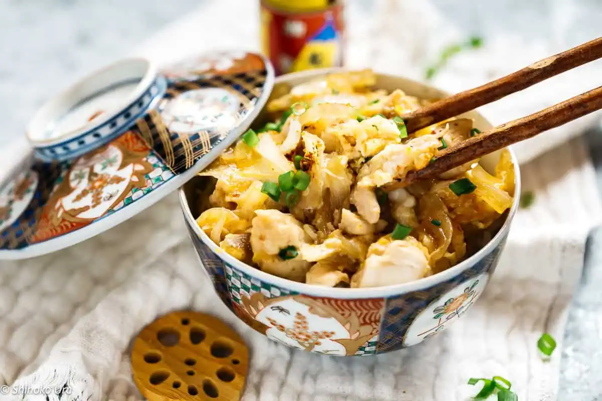 oyako don served in a donburi bowl with a lid and a pair of chopsticks digging into the oyakodon