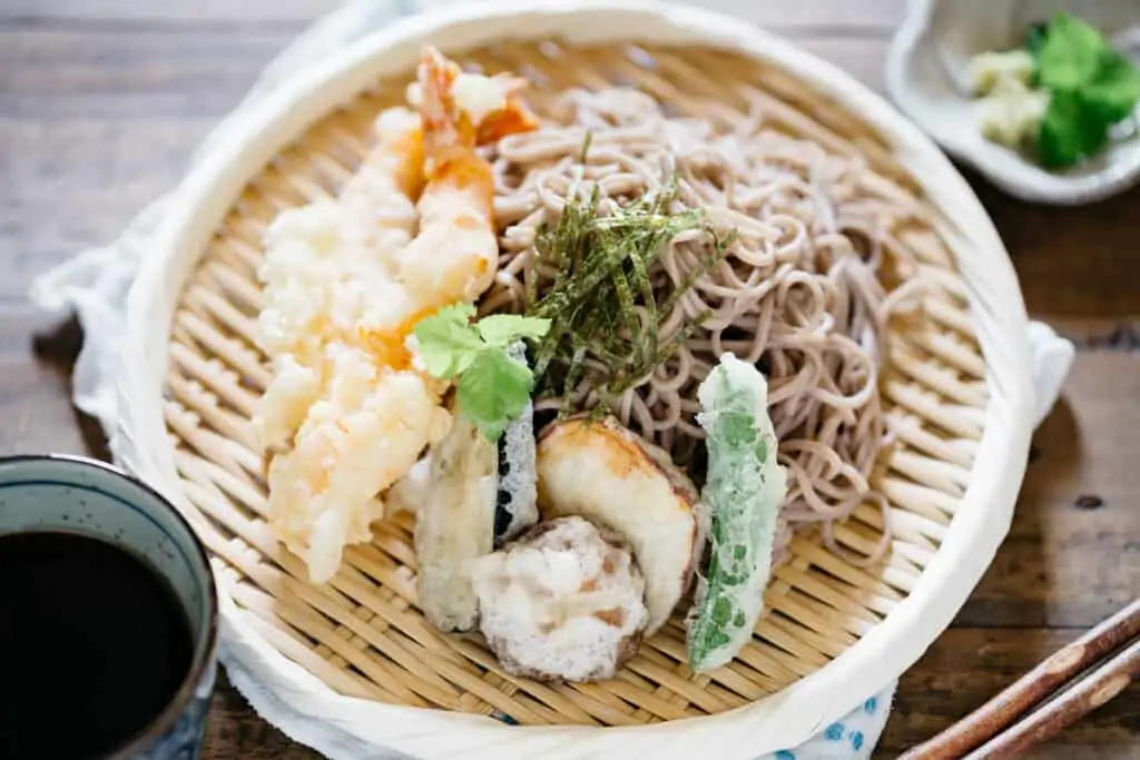 Tempura pieces and soba noodles served on a bamboo tray with dipping sauce in a small cup