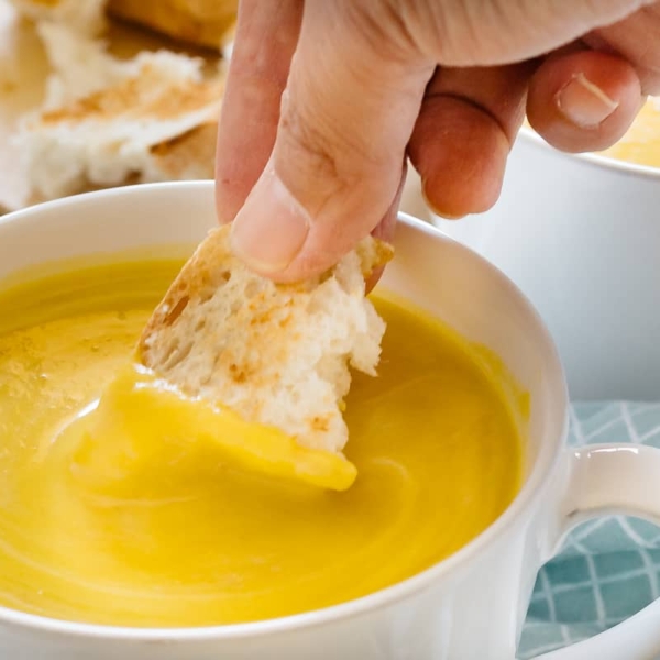 A piece of bread in a hand dipped in kabocha squash soup in a white soup mug
