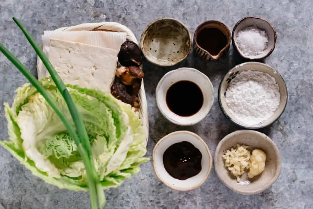 cabbage leaves, scallions, firm tofu, three shiitake mushrooms on the bamboo tray, condiments in small bowls 