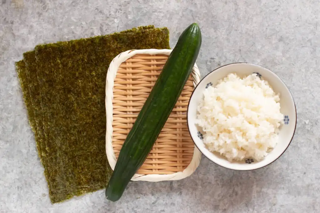 three nori sheets, cucumber on a bamboo tray, and a bowl of sushi rice 