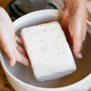 a block of tofu transported into a bowl of cold water by two hands