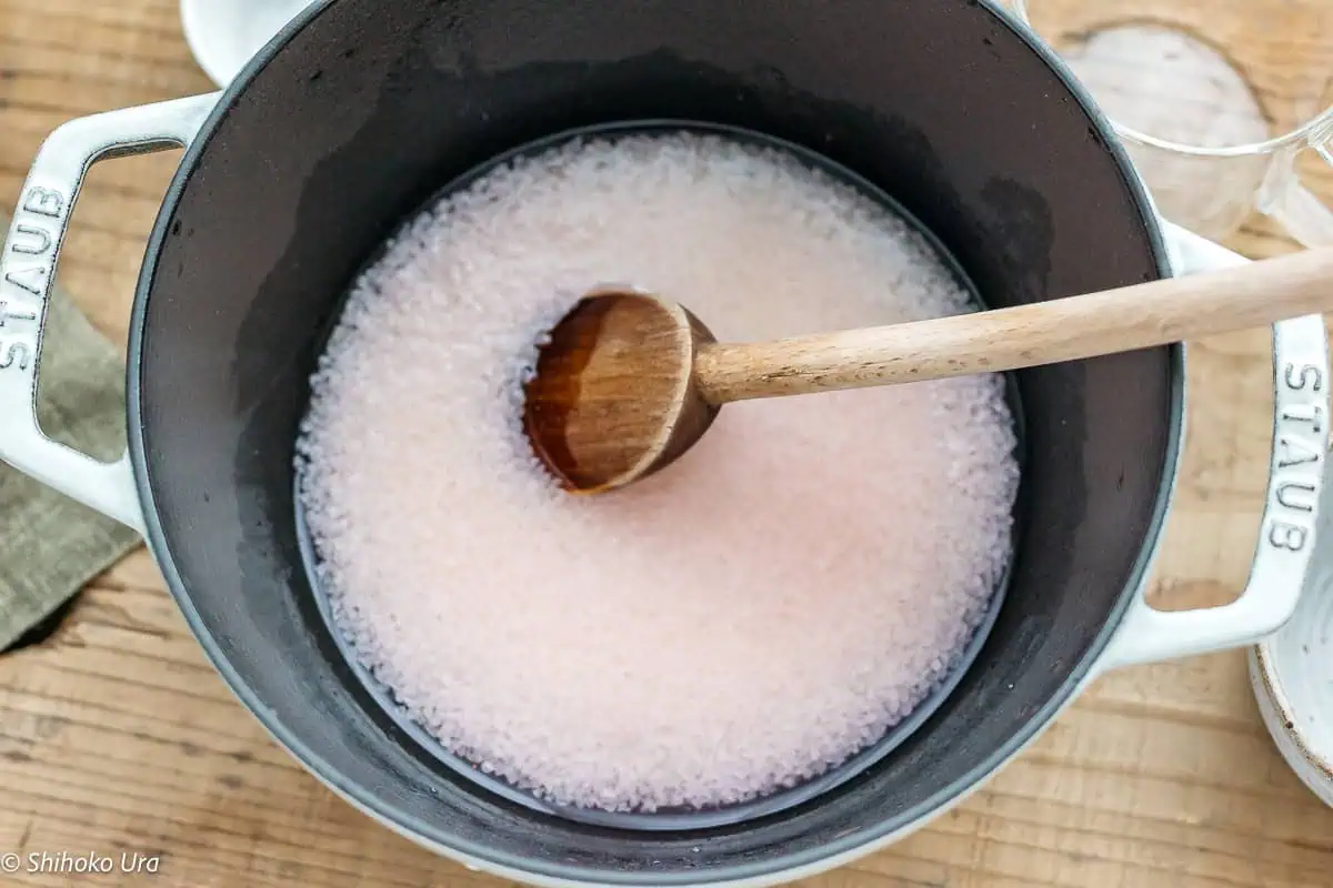 pink coloured water, sugar, and domyojiko in a pot with a wooden spatula