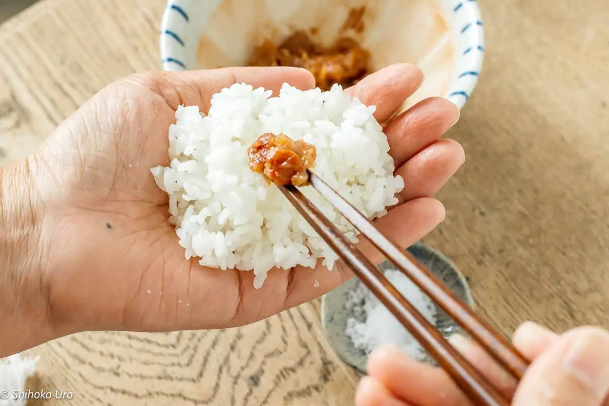 Hand holding rice ball with umeboshi filling, chopsticks ready to eat.